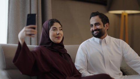 تعابير الموافقة و القبول، الأجواء العائلية السعيدة، توثيق اللحظات السعيدة ، تقوية العلاقات الزوجية وروابط الحب والمودة، التقاط صور السيلفي التذكارية باستخدام الهاتف المحمول، زوجان عربيان خليجيان سعوديان يرتديان الزي التقليدي يجلسان فى  غرفه المعيشة يستخدمان الجوال