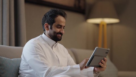 Using a modern advanced technical device, performing tasks from home, working remotely, an Arab Gulf Saudi man wearing a white thobe sitting in the living room working on a tablet, integrating technology and tech into daily life, looking at the camera with expressions of happiness and joy.