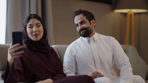 مفهوم الأمان والإستقرار العائلي، إجراء مكالمة فيديو أونلاين، توثيق اللحظات العائلية السعيدة، زوجين عربيين خليجيين سعوديين يجلسان فى غرفه المعيشة يستخدمان الهاتف المحمول، المحافظة على الترابط الأسري، ايماءات السعادة و السرور