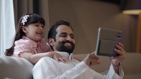 Parents' interest in their children, integrating learning with technology, following online educational platforms, watching recreational and educational activities, an Arab Gulf Saudi man wearing a white thobe sitting with his daughter and using a tablet, gestures of happiness and joy.
