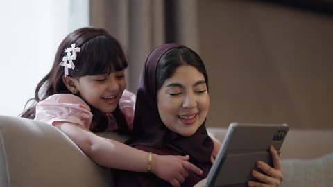 دمج التعليم مع التقنية والتكنولوجيا، متابعة منصات التعليم الالكتروني المتاحة عبر الانترنت بإستخدام التابلت، حل الواجبات المنزليه ، أم عربية خليجة سعودية تجلس فى غرفة المعيشة تقوم بتعليم وتدريس ابنتها