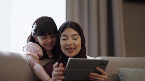 متابعة منصات التعليم الالكتروني المتاحة عبر الانترنت بإستخدام التابلت، دمج التعليم مع التقنية والتكنولوجيا، حل الواجبات المنزليه، ، أم عربية خليجة سعودية تجلس فى غرفة المعيشة تقوم بتعليم وتدريس ابنتها