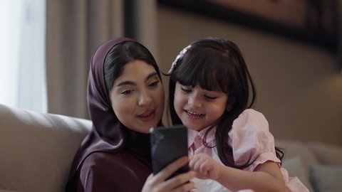 The concept of motherhood and childhood, spending enjoyable times with children, making a video call, an Arab Gulf Saudi woman sitting with her daughter taking a commemorative selfie using a mobile phone, the mother's care and attention to her children, family love and affection.