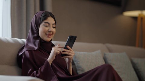 الدردشة والتواصل مع التواصل مع الأصدقاء اونلاين، متابعة أخبار وسائل التواصل الاجتماعي باستخدام الجوال، إمرأة عربية خليجية سعودية مبتسمة ترتدي حجاب وعباءة تجلس على الاريكة تمسك بيديها الهاتف المحمول، تعابير وايماءات الفرح والسرور
