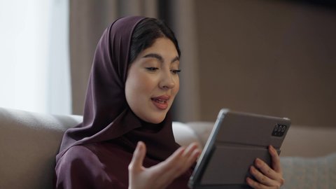 الدردشات والمحادثات عبر مكالمات الفيديو, تبادل الأحاديث المسليه عبر التابلت، الدردشة و التواصل الاجتماعي, ايماءات السعادة والسرور، تصوير مقرب لامراه عربيه خليجيه سعودية محجبة ترتدي العباءة تجلس فى غرفه المعيشة تقوم باستخدام الجهاز اللوحي