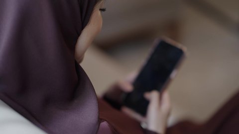 التواصل مع الأصدقاء عبر المنصات الإلكترونية، تصفح وسائل التواصل الاجتماعي باستخدام الجوال،  تصوير مقرب من الخلف لامراة عربية خليجية سعودية محجبة ترتدي العباءة و الحجاب تمسك بيديها الهاتف المحمول