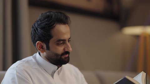 تنمية مهارات القراءة، قضاء أوقات الفراغ بالتثقيف والتعلم, الاهتمام بتصفح الكتب المفيدة، تصوير مقرب لرجل عربي خليجي سعودي يرتدي الثوب التقليدي يجلس فى غرفة المعيشة ويحمل بيده كتاب