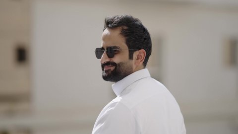 استخدام العدسات الطبية الملونة لحماية العين من أشعة الشمس، الاهتمام بالأناقة والمظهر الخارجي، مفهوم الرجولة والثقة بالنفس، تصوير مقرب لرجل عربي خليجي سعودي مبتسم يرتدي الثوب الابيض والنظارة الشمسية ينظر الى الكاميرا بإيماءات السرور