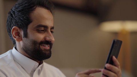 Making a video call via mobile, communicating with family and friends during free time, a close-up shot of a Gulf Arab Saudi man wearing traditional attire and holding a mobile phone in his hand, gestures of happiness and joy.
