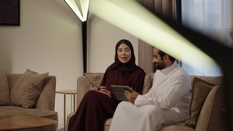 إنجاز المهام في المنزل، ادارة الأعمال عن بعد، المشاركة والتعاون وتبادل الآراء بين الزوجين، استخدام الأجهزة والتقنيات الحديثة والمتطورة،رجل عربي خليجي سعودي يرتدي الثوب الابيض يقوم باستخدام جهاز التابليت اللوحي بجانب زوجته