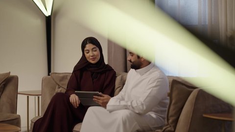 ادارة الأعمال عن بعد، المشاركة والتعاون وتبادل الآراء بين الزوجين، استخدام الأجهزة والتقنيات الحديثة والمتطورة،رجل عربي خليجي سعودي يرتدي الثوب الابيض يقوم باستخدام جهاز التابليت اللوحي بجانب زوجته، إنجاز المهام في المنزل