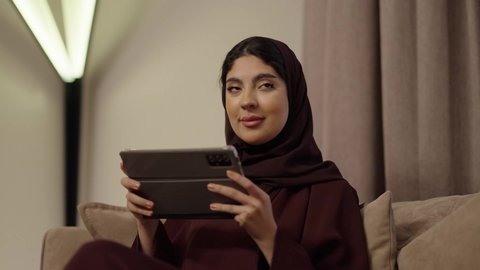 Browsing the internet and social media through a tablet, celebrating the anniversary of the wedding day, an Arab Gulf Saudi man wearing a white thobe presents a gift in a box to his wife on the occasion of Valentine's Day, expressing intimacy and love between the couple, gestures of happiness and joy.