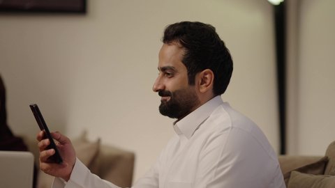 تصفح الانترنت ومواقع التواصل الاجتماعي عبر الجوال، تبادل الاحاديث والاخبار بين افراد العائلة، المحافظة على الترابط الأسري، مفهوم الأمان والإستقرار العائلي، امراه عربيه خليجيه سعودية ترتدي العباءة و الحجاب تجلس فى غرفه المعيشة برفقة زوجها تستخدم الحاسب المحمول