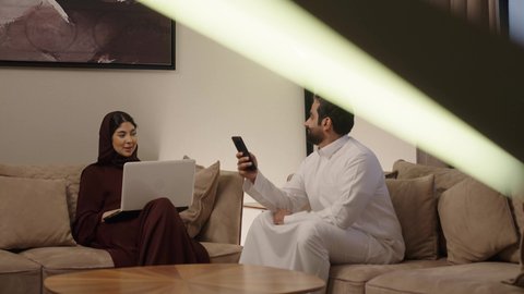 تبادل الاحاديث والاخبار بين افراد العائلة، المحافظة على الترابط الأسري، مفهوم الأمان والإستقرار العائلي، زوجين عربيين خليجيين سعوديين يجلسان على اريكة مريحة في غرفة المعيشة يستخدمان الهاتف المحمول و الابتوب