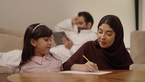 مفهوم العودة إلى المدارس، مراجعة الدروس اليومية، التعليم في المملكة العربية السعودية, الاستعداد للعام الدراسي الجديد,فتاة عربية خليجية سعودية ترتدي الزي الكاجوال تجلس فى غرفة المعيشة برفقة والديها, اهتمام الآباء بالأبناء، استخدام جهاز التابلت