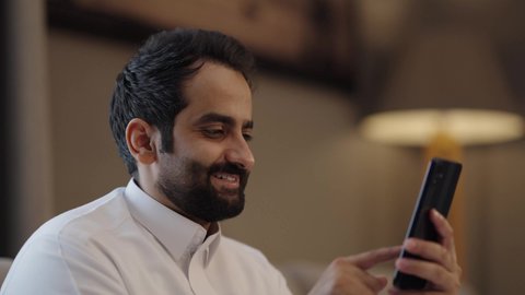 Browsing social media, an Arab Gulf Saudi man wearing traditional attire holds a mobile phone in his hand, chatting and communicating with family and friends, using a modern and advanced technological device, working remotely and completing required tasks, gestures of happiness and joy.