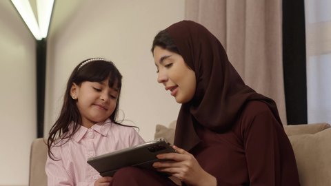 تعابير الموافقة و القبول، اهتمام الآباء بالأبناء، دمج التعلم بالتكنولوجيا، متابعة منصات التعليم الالكتروني عبر الانترنت، مشاهدة الأنشطة الترفيهية والتعليمية، فتاه عربية خليجية سعودية ترتدي لباس الكاجوال تستخدم التابلت تجلس برفقة والدتها