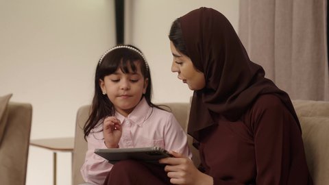 اهتمام الآباء بالأبناء، دمج التعلم بالتكنولوجيا، متابعة منصات التعليم الالكتروني عبر الانترنت، مشاهدة الأنشطة الترفيهية والتعليمية، فتاه عربية خليجية سعودية ترتدي لباس الكاجوال تستخدم التابلت تجلس برفقة والدتها
