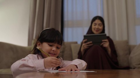 Developing and improving writing skills, diligence and preparation for school tests, a Saudi Arabian Gulf girl is doing her homework, an Arab Gulf woman in Saudi Arabia wearing an abaya and hijab is sitting on the couch holding a tablet in her hands.