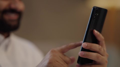 Browsing social media, a close-up shot of the hand of an Arab Gulf Saudi man holding a mobile phone, chatting and communicating with family and friends, using a modern and advanced technological device, working remotely and completing required tasks, tapping on the mobile screen.