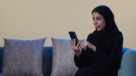 Providing home care services, women's professions and jobs, a portrait of a foreign cleaning worker cleaning the house, an Arab Gulf Saudi woman wearing a hijab and an abaya sitting on the sofa holding a mobile phone, browsing social media, brown background.