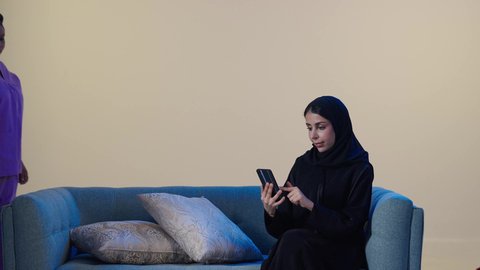 Trained and qualified domestic workers, women's professions and jobs, a portrait of a foreign cleaning worker cleaning the house, an Arab Gulf Saudi woman wearing a hijab and an abaya sitting on the sofa holding a mobile phone, browsing social media, brown background.