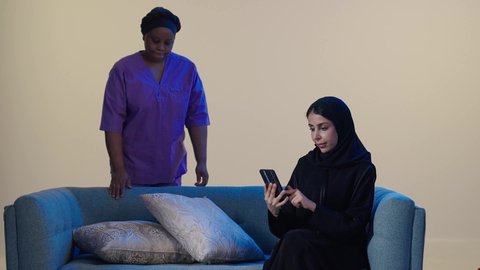 Women's professions and jobs, trained and qualified domestic helpers, a portrait of a foreign cleaning worker cleaning the house, an Arab Gulf Saudi woman wearing a hijab and an abaya sitting on the sofa holding a mobile phone, browsing social media, brown background.
