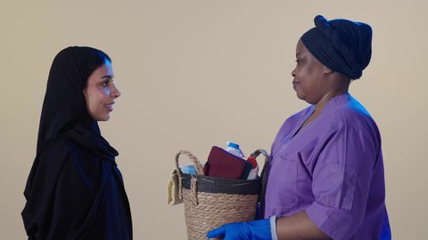 النظر الى الكاميرا بتعابير السعادة و السرور، الاهتمام بالتنظيف والقيام بالمهام الروتينية، عاملة نظافة أجنبية تحمل بيدها سلة قش بأدوات تنظيف المنزل تقف برفقة ربة المنزل ، مهن ووظائف نسائية، خلفية بنية