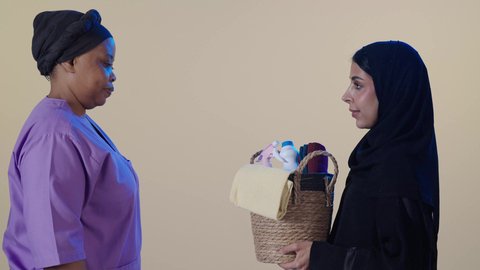 الاهتمام بالتنظيف والقيام بالمهام الروتينية، عاملة نظافة أجنبية تحمل بيدها سلة قش بأدوات تنظيف المنزل، بورتريه لربة منزل عربية خليجية سعودية محجبة  تعطي ادوات التنظيف  للخادمة الإفريقية، مهن ووظائف نسائية، خلفية بنية