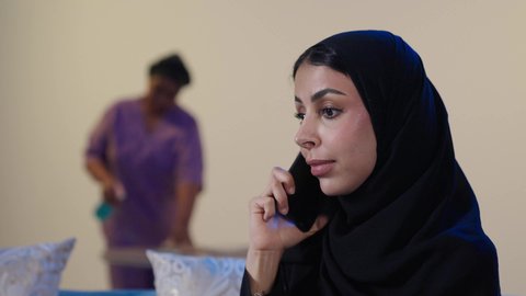 المهام والأعمال المنزلية الروتينية، عاملة نظافة ترتدي اليونيفورم الخاص بالعمل تقوم بمسك وتنظيف البيت، مهن ووظائف نسائية، جودة الخدمات في السعودية، تصوير مقرب لامراه عربيه خليجيه سعودية ترتدي العباءة و الحجاب تقوم باجراء مكالمة هاتفية عبر الجوال