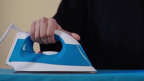 Completing tasks and routine work, using electrical appliances, drying and organizing clothes, a close-up portrait of a Saudi Gulf Arab woman wearing a black abaya ironing clothes with an iron, the concept of household management, brown background.