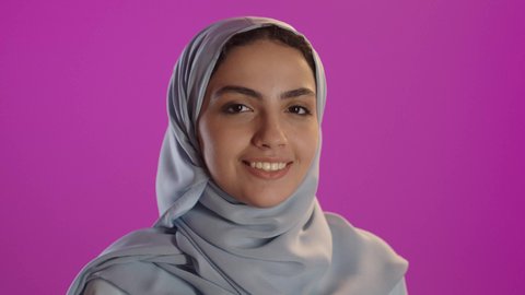 Elegant and modern hijab fashion, authentic Arab features, the concept of self-confidence and attention to appearance, a close-up portrait of a smiling Saudi Arabian Gulf woman wearing a hijab and abaya, looking at the camera with expressions of joy, standing with her hands crossed, purple background