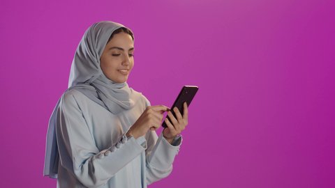 Enjoying time on her mobile phone with friends; a portrait of a veiled Saudi Arabian woman wearing an abaya and using a mobile phone, showcasing modern technology; purple background.