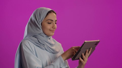Connecting with family and friends via technology; a portrait of a veiled Saudi Arabian woman wearing a grey abaya, holding a tablet and browsing social media; purple background.