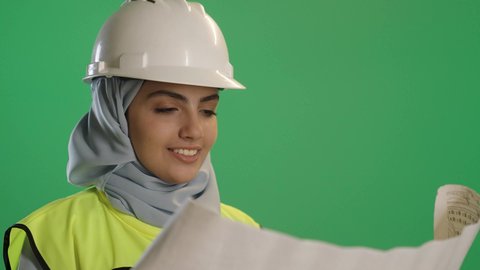 الشغف للوصول إلى النجاح وتحقيق الأحلام , بورتريه مقرب لمهندسة عربية خليجية سعودية مبتسمة ترتدي سترة العمل تمسك بيدها مخططات هندسية وتقف بإيماءات الثقة والفخر,الهندسة المعمارية ومخططات الانشاء, خلفية خضراء كروما
