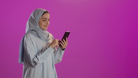 Browsing social media, a portrait of a veiled Saudi Arabian woman wearing an abaya, using a mobile phone, utilizing modern technology, spending enjoyable time on her phone with friends, purple background.