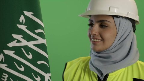 السعادة بالانتماء للهوية السعودية, اليوم الوطني السعودي 23 سبتمبر, بورتريه مقرب لمهندسة عربية خليجية سعودية ترتدي سترة الحماية والخوذة,  تمسك علم المملكة بإيماءات الفخر, يوم العلم 11 مارس, مهن ووظائف نسائية،, خلفية خضراء, كروما
