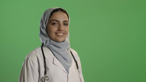 مفهوم الطب والرعاية الصحية، العمل في القطاع الصحي في المملكة،بورتريه لطبيبة عربية خليجية سعودية ترتدي العباءة والحجاب مع المعطف الابيض تقف باستقامة، النظر بايماءات السعادة والسرور، مهن ووظائف نسائية، تقديم الخدمات للمرضى بكل كفاءه، خلفيه خضراء, كروما
