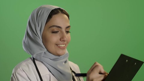 تقديم الخدمات والاستشارات الطبية للمراجعين، بورتريه مقرب لطبيبة عربية خليجية سعودية ترتدي  المعطف الأبيض و السماعة تحمل بيدها دفتر تدوين المعلومات العامة عن حالة المريض، مهن ووظائف نسائية، دراسة و مراجعة حالات المرضى , خلفية خضراء, كروما
