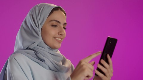 Using modern technology, sharing happy moments on social media, spending enjoyable time on mobile with friends, a portrait of a veiled Saudi Arabian woman wearing a gray abaya using a mobile phone, purple background