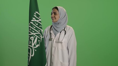 اليوم الوطني السعودي 23 سبتمبر, بورتريه لطبيبة عربية خليجية سعودية ترتدي الحجاب مع المعطف الابيض, النظرالى علم المملكة بإيماءات الفخر, التباهي بإنجازات البلاد, خلفية خضراء, كروما