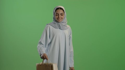 إيماءات تدل على السعاده، عروض وتخفيضات موسميه، الاستمتاع بالتبضع فى السعودية، التسوق في المراكز التجاريه، بورتريه لإمرأة عربيه خليجية سعوديه محجبة ترتدي عباءه رمادية تحمل بيدها أكياس التسوق، خلفية خضراء, كروما