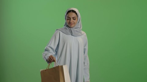 عروض وتخفيضات موسميه, إيماءات تدل على السعاده، الاستمتاع بالتبضع فى السعودية, التسوق في المراكز التجاريه، بورتريه لإمرأة عربيه خليجية سعوديه محجبة ترتدي عباءه رمادية تحمل بيدها أكياس ملونة، خلفية خضراء, كروما