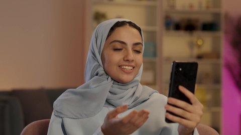 التواصل مع الأقارب والأصدقاء باستخدام الاجهزة التقنية الحديثة، امرأة عربية خليجية سعودية محجبة ترتدي العباءة الرمادية تقوم بإستخدام الهاتف المحمول، الجلوس في غرفة المعيشه بإيماءات السرور والسعادة