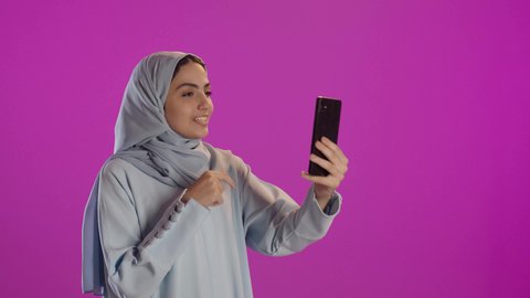 مشاركة اللحظات السعيدة على وسائل التواصل الاجتماعي, قضاء وقت ممتع عبر الجوال مع الأصحاب، بورتريه لامرأة عربية خليجية سعودية محجبة ترتدي العباءة الرمادية تقوم بإستخدام الهاتف المحمول، خلفية بنفسجية