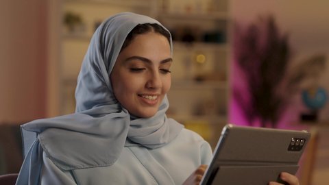 استخدام التكنولوجيا الحديثة، امرأة عربية خليجية سعودية محجبة ترتدي العباءة الرمادية تستخدم التابلت وتقوم بتصفح وسائل التواصل الإجتماعي, الجلوس في غرفة المعيشه بإيماءات السرور والسعاده