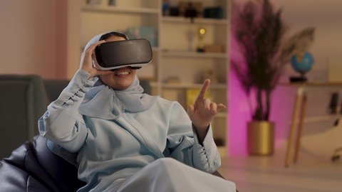 محاكاة العالم الخيال, امراءة عربية خليجية سعودية ترتدي الحجاب والعباءة ونظارات الواقع الافتراضي تجلس على الكرسي تقوم بمد يديها الى الامام ومحاكاة العالم الخيالي, محاولة لمس شيء ما،