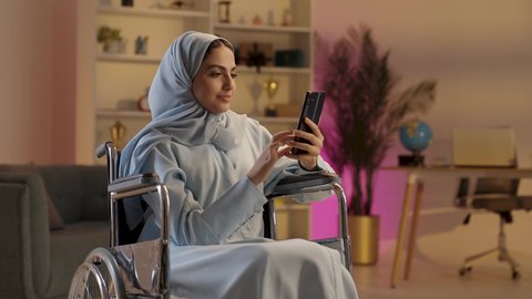 سيده عربيه خليجيه سعوديه محجبة ترتدي العباءة الرمادية تجلس على كرسي متحرك من ذوي أصحاب الهمم تقوم بإستخدام الهاتف المحمول, مشاركة اللحظات السعيدة على وسائل التواصل الاجتماعي, قضاء وقت ممتع عبر الجوال مع الأصحاب