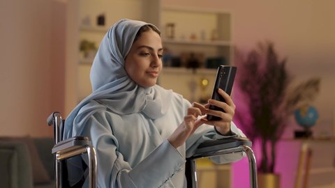 إنجاز المشاريع ومهام العمل عن بعد، التواصل مع الأهل والأصدقاء عبرالجوال، سيدة عربيه خليجيه سعوديه من ذوي الاحتياجات الخاصة ترتدي العباءة الرمادية والحجاب تجلس على كرسي متحرك تستخدم الهاتف