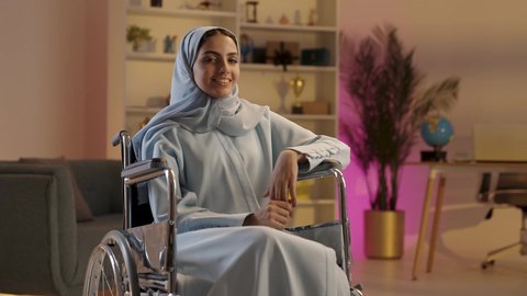 الجلوس في غرفة المعيشه بإيماءات السرور والسعادة, سيدة عربيه خليجيه سعوديه من ذوي الاحتياجات الخاصة ترتدي العباءة الرمادية والحجاب تجلس على كرسي متحرك تنظر إلى الكاميرا